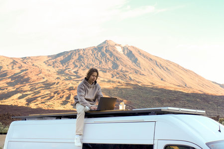 A young man is seen using a laptop while sitting on the roof of a van with the majestic Teide...