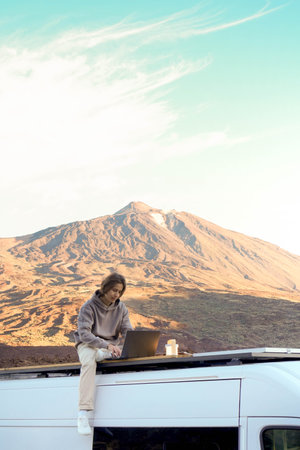 Man sits on a roof with a laptop in front of a majestic mountainous landscape.
