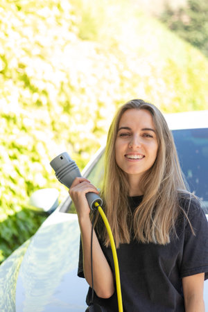 Vertical view of young beautiful woman smiling holding an electric car plug in green background as renewable energy conceptの写真素材