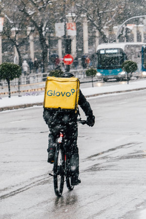 Madrid, Spain - January 2021: Glovo (food delivery company) rider in Alacala street with hevy snow due to Filomena Storm. bus in the background. View from the back. Rider on bike.のeditorial素材