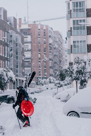 Madrid, Spain - January 2021: a person with skis and a sled in a street in the capital of Spain with heavy snow due to Filomena Storm. Red sled and long street with buildings.のeditorial素材