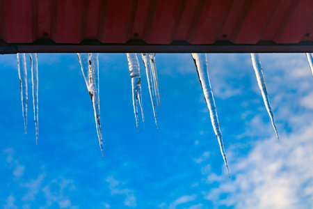 beautiful icicles shine in the sun against the blue sky. landscape with ice icicles hanging from the roof of the house.の写真素材
