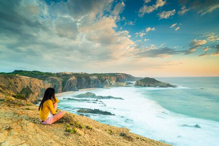 27/09-18, Zambujeira do Mar, Portugal. A girl is sitting by the edge of a cliff. Big waves are crashing on the beach below.の写真素材