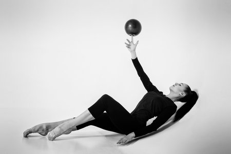 a girl from rhythmic gymnastics in a bodysuit shows lying on her back twirling a ball on the finger of her raised handの写真素材