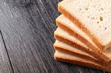 Slices of freshly baked bread are neatly stacked on a dark wooden surface, highlighting their light...