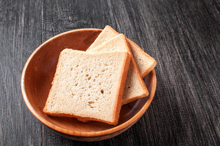 Three slices of fresh bread arranged neatly in a round wooden bowl on a rustic dark wooden table,...