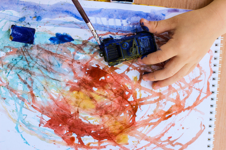 Top view of watercolor container, album with abstract preschooler's drawings and child's hand holding toy auto.の写真素材