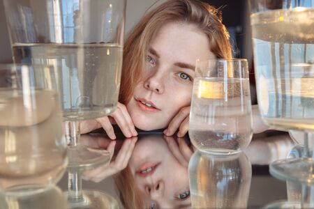 A blonde with cute features looks into the frame. The girl is reflected in the mirror. The girl is surrounded by water containers. the light is falling on the girl from behind.の写真素材