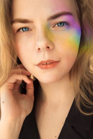 Portrait of a blue-eyed girl. She has a milky facial features. A rainbow of saturated colors falls on the face. A hand reaches for the neck. The girl has blond hair.の写真素材