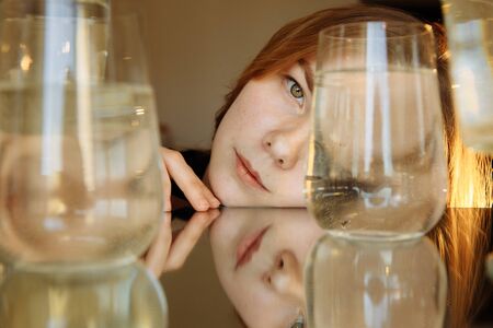 The blonde girl is looking into the frame. One eye is closed by a container of water. Man is reflected in the mirror. Everywhere there are containers with water. The girl is fresh and young.の写真素材