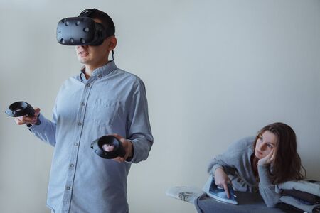 A man enthusiastically plays video games in virtual reality. The wife is doing household chores at this time. High quality photoの写真素材