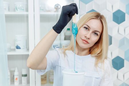 Master of sugaring. The blonde holds a wooden spatula with blue wax. The wax drips from the spatula into the jar. The girl has big eyes and beautiful features. The woman has a smile on her face.の写真素材