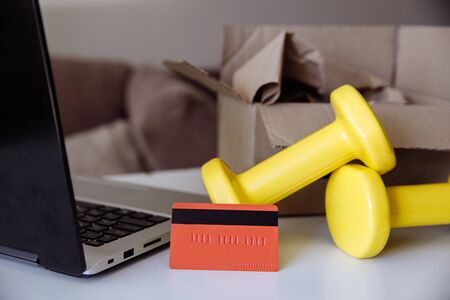 On a white table are: a black laptop, a red Bank card, yellow dumbbells, and a box. The dumbbells have just been taken out of the box. Purchase of sporting goods in an online store with delivery.の写真素材