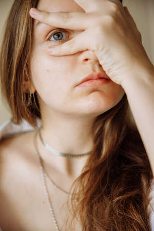 The girl looks at the camera through her fingers. Tender beautiful portrait of a brunette. The brunette has gray eyes. The woman has several ornaments around her neckの写真素材