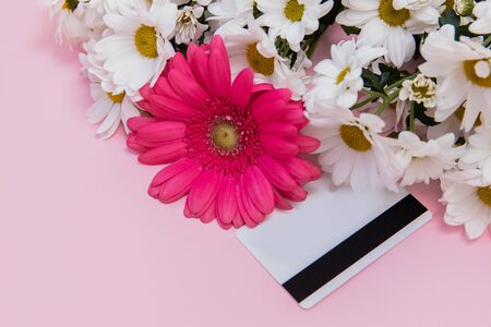 Pink gerbera and white chamomile flowers are on the pink table. Next to the flowers is a white plastic Bank card. There is space for text.の写真素材