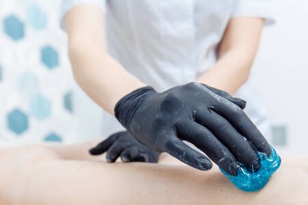 Hand in a black glove gets soft blue wax for hair removal on the leg. There are hairs on the leg. A master in a white coat is seen in the background.の写真素材