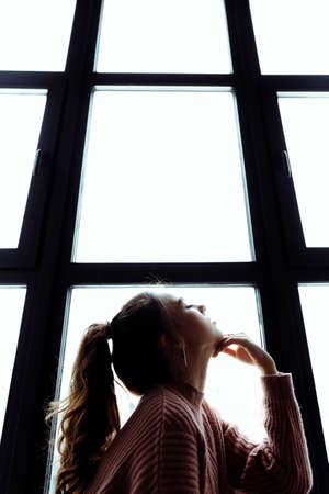 a brunette girl is sitting opposite the window. The girls hair is pulled back in a ponytail. The window looks like a grid. The girl is wearing a pink sweater. High quality photoの写真素材