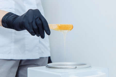 Yellow beeswax off with a wooden spatula for waxing. The spatula is held by the hand of a man in a black latex glove. The wax flows into a special bath. High quality photoの写真素材