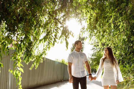 A man in a white shirt and a girl in a white dress are walking forward holding hands. Young people can be seen through the trees. The sun is shining in my back. High quality photoの写真素材
