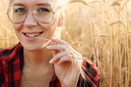 A woman with gray eyes wearing glasses and a red checked shirt is sitting in a field. The woman clamped her beautiful teeth on an ear of wheat. High quality photoの写真素材