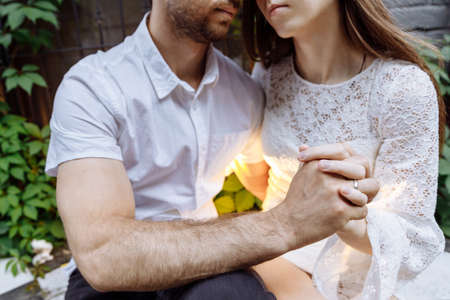 A man and a woman in white clothes embrace. Peoples hands are intertwined. The mans lips are drawn...