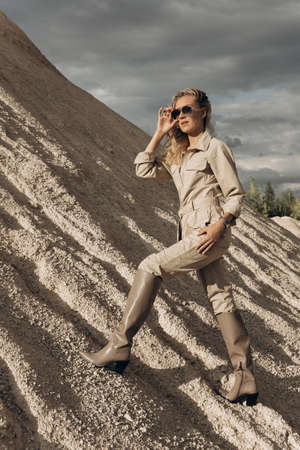 A tall young woman stands in a sand dune environment. The blondes gaze is directed to the side. Dark...
