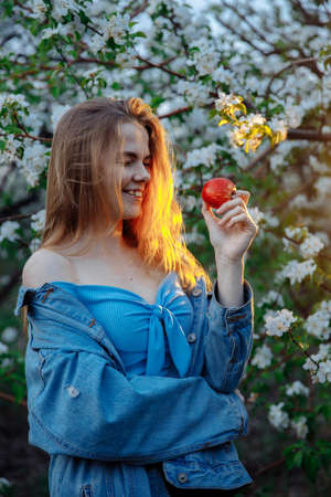 Young woman in denim jacket smiling while looking at an apple. The blonde hair glows with warm light. In the background a blooming garden. High quality photoの写真素材