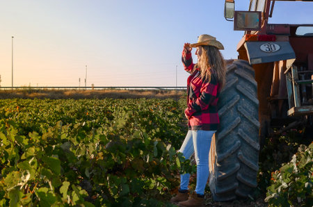 farming woman, looking at the horizon, in a vineyard field with a red tractor behind, a straw hat and red plaid shirt.の写真素材