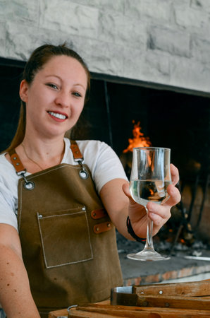 Smiling woman toasting with a glass of white wine in her hand, next to a grill wearing a brown apron and white t-shirtの写真素材