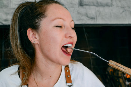 Smiling woman with light eyes, eating lamb meat, next to a grill in the foreground