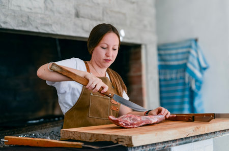 Smiling woman preparing to cook a roast, with a leg of lamb on a wooden board, next to a griller, wearing a brown apron and white t-shirt, using kitchen utensilsの写真素材