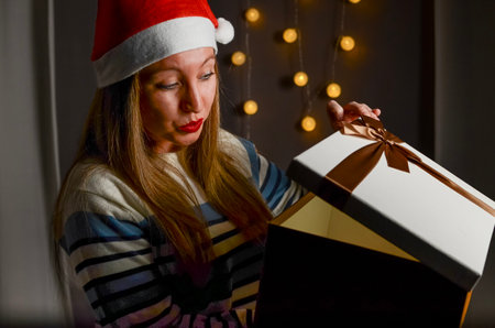 Woman opening her Christmas present wearing a red Santa hat and a striped sweater, and Christmas...
