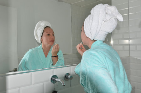Woman wearing bathrobe and towel on her head applying lip gloss while looking in the bathroom mirrorの写真素材