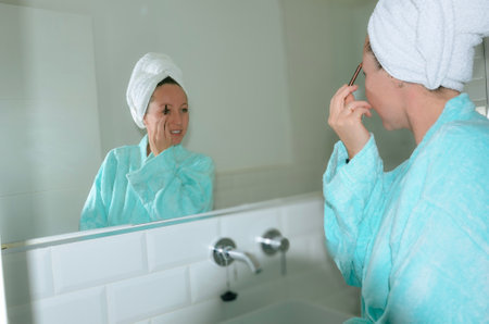 Woman wearing bathrobe and towel puts on makeup in front of a bathroom mirrorの写真素材