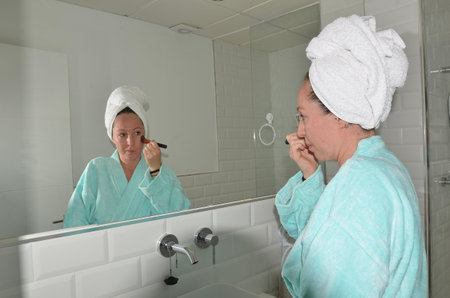 Woman applying makeup with brush in bathroom mirror wearing bathrobe and towel turbanの写真素材