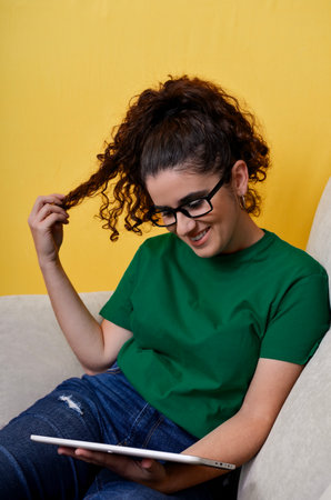 Happy casual student using digital tablet on sofa and playing with hair