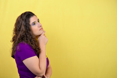 Young woman with curly hair and purple t-shirt is thinking with hand on chin, looking up on yellow backgroundの写真素材