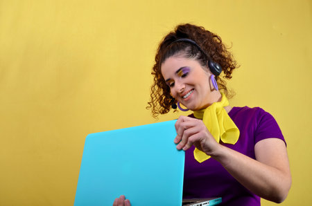 Happy female telemarketer opening her light blue laptop while wearing a headset and smiling on a yellow backgroundの写真素材