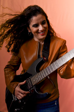 Female musician with long, curly hair, wearing a brown leather jacket, passionately playing a black electric guitarの写真素材