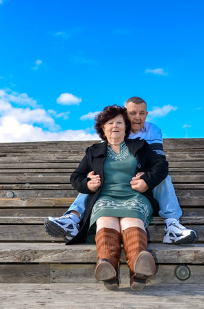 Happy retired couple holding hands and smiling, sitting on wooden steps under a blue skyの写真素材
