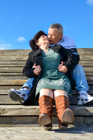 Senior couple enjoying a romantic moment, kissing on outdoor wooden steps under a clear blue skyの写真素材
