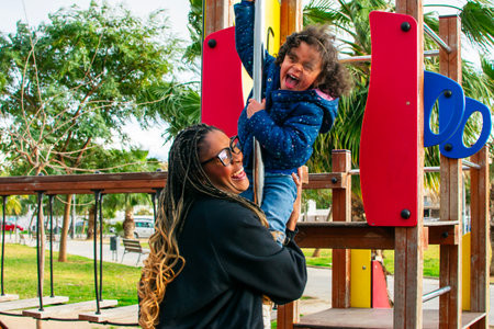Mother supporting her daughter while playing on a playground slide in an urban park, enjoying quality time togetherの写真素材