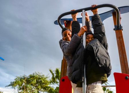 Two siblings are hanging on a playground structure, enjoying a sunny day at the park