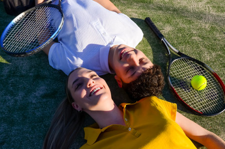 Two young tennis players are lying on the court with their rackets and a tennis ball, enjoying the sunshine after a matchの写真素材