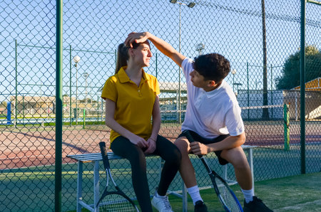 Two young tennis players are sitting on a bench, taking a break after a tennis match, with the boy gently touching the girl's foreheadの写真素材