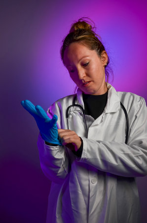 Doctor putting on blue medical gloves, wearing white coat and stethoscope, with purple and blue backgroundの写真素材