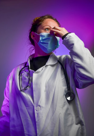 Female doctor wearing surgical mask and white coat with stethoscope, touching her face under purple lightの写真素材