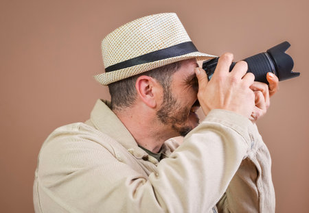Smiling photographer wearing beige hat taking photos with professional camera in studio with brown backgroundの写真素材