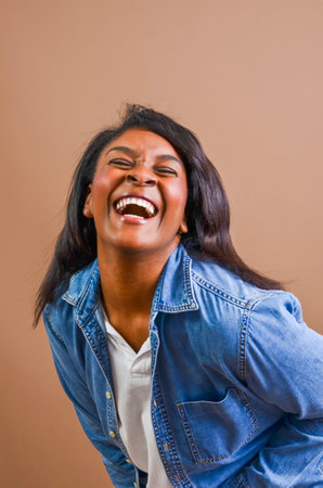 Studio portrait of a cheerful african american woman with long black hair, wearing a denim shirt and white t-shirt, expressing pure joy and laughterの写真素材