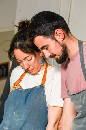 Two artisans wearing aprons are working together, focusing their attention on a pottery project in...